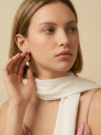 Close-up of sculptural gold link earrings featuring a twisted rope texture and hanging pink freshwater baroque pearl drops. The earrings are displayed on a champagne silk fabric to highlight the pearl luster and high-shine gold finish
