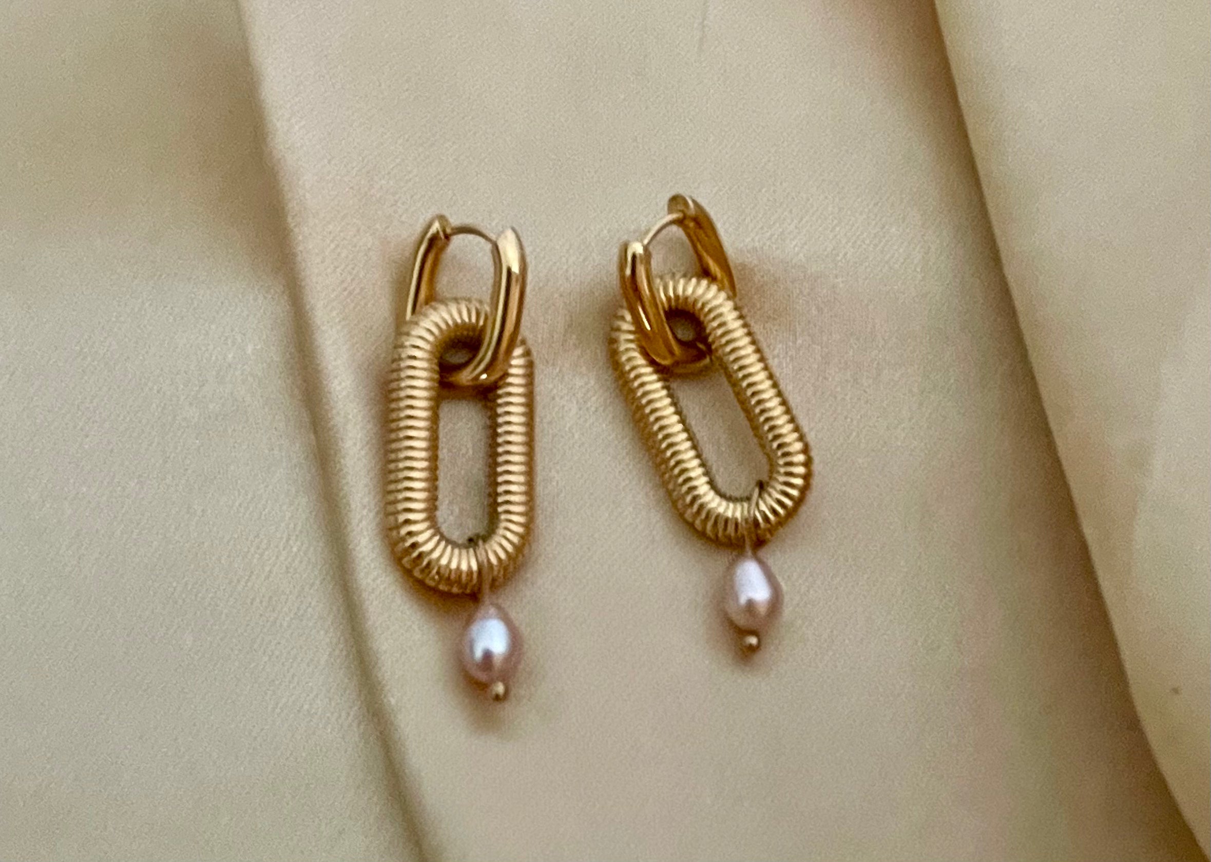 Close-up of textured gold oval link earrings featuring a dainty pink pearl drop. High-resolution commercial jewelry photography showcasing ribbed gold detailing and lustrous pearl against a soft matte ivory background.
