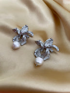 Close-up of sculptural silver orchid earrings featuring a high-luster real white pearl drop. The textured metal petals mimic a blooming iris flower, perfect for bridal jewelry or statement wear