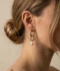 Close-up of sculptural gold link earrings featuring a twisted rope texture and hanging freshwater baroque pearl drops. The earrings are displayed on a champagne silk fabric to highlight the pearl luster and high-shine gold finish