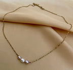 Close-up of a model wearing a dainty gold chain necklace featuring a trio of small pearls: a central white pearl flanked by two pink oval baroque pearls, styled with an ivory silk top