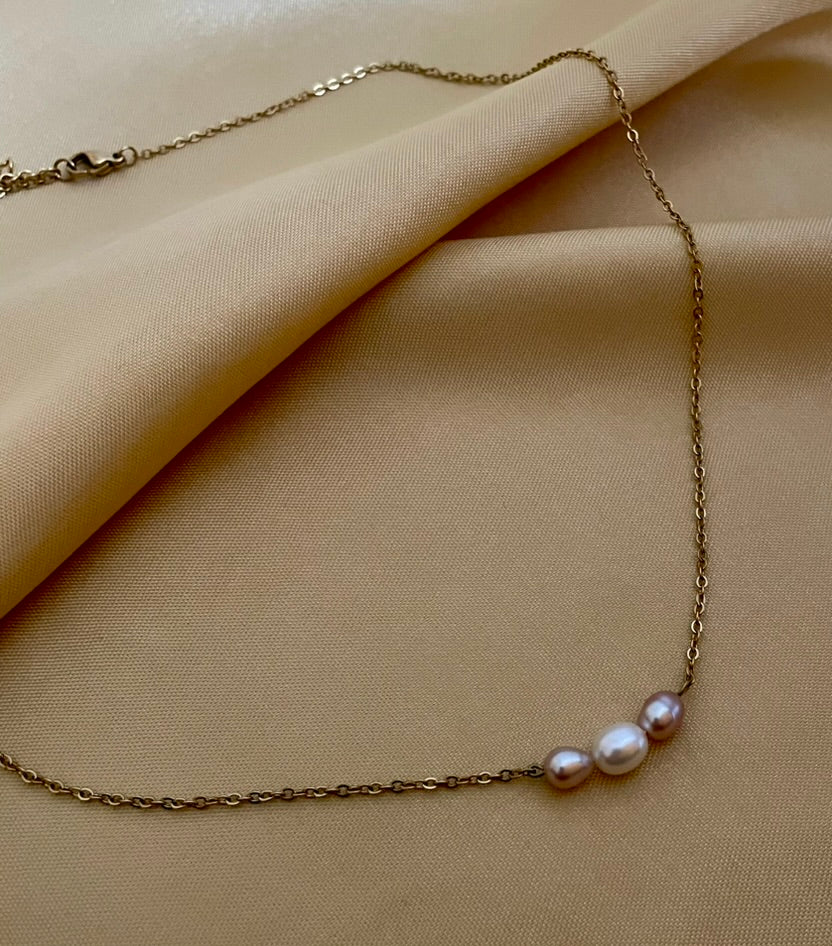 Close-up of a model wearing a dainty gold chain necklace featuring a trio of small pearls: a central white pearl flanked by two pink oval baroque pearls