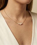 Close-up of a model wearing a dainty gold chain necklace featuring a trio of small pearls: a central white pearl flanked by two pink oval baroque pearls, styled with an ivory silk top