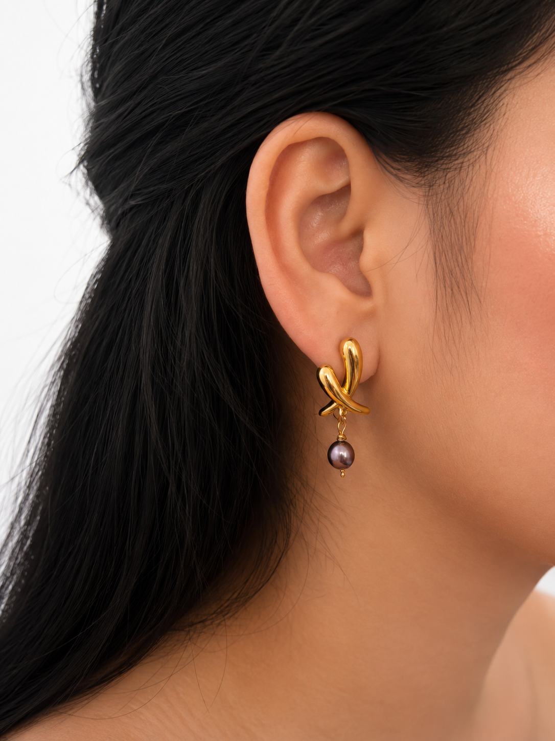 Close-up of a gold cross stud earring with a dark baroque freshwater pearl drop worn on the ear