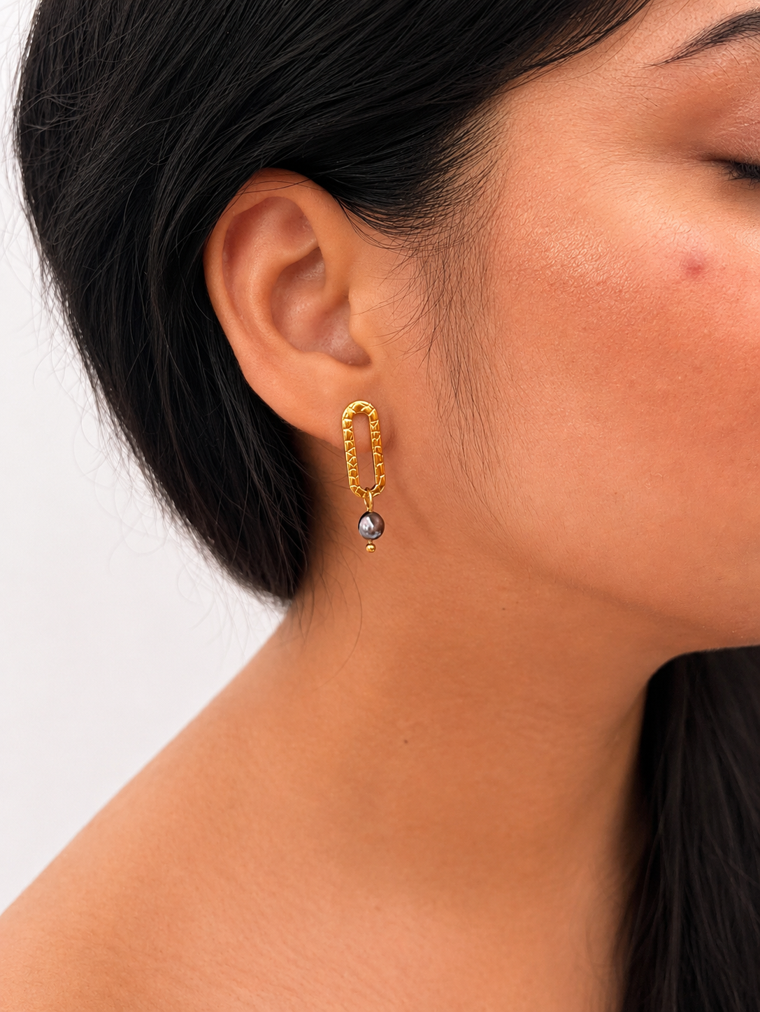 Close-up of a textured gold stud earring with a 6 mm dark freshwater pearl worn on the ear.