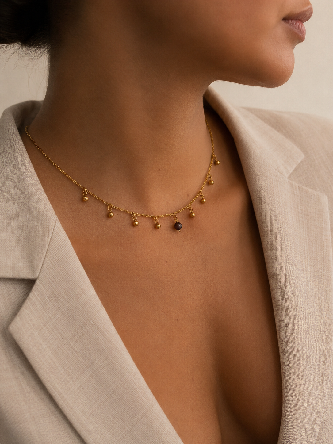Woman wearing a gold charm necklace with a dark grey freshwater pearl and suspended gold bead accents.