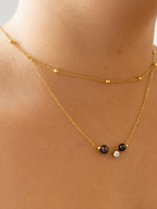 Close-up of a gold layered necklace with two dark freshwater pearls and a cubic zirconia pendant worn on the neck