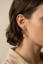 Close-up editorial profile of a model wearing detailed gold openwork hoop earrings featuring a leaf motif and a single dangling white oval pearl. The warm, soft lighting highlights the pearl's luster and the gold texture against the model's skin, set against a neutral background