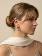 Close-up of 18k gold multi-hoop earrings featuring a lustrous grey pearl drop, worn by a model with a low bun hairstyle. The jewelry highlights a textured gold finish and organic pearl shape, set against a plain ivory background
