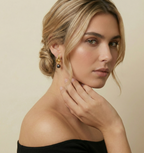 Close-up portrait of a female model wearing elegant gold twisted hoop earrings featuring a lustrous black pearl drop. She has a sophisticated updo hairstyle and wears a black off-shoulder top, highlighting the jewelry against a neutral background