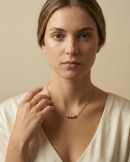 Close-up editorial portrait of a French model wearing a delicate gold chain necklace featuring three small, oval pink pearls with a high silvery luster, styled with a deep-neck ivory silk top against a matte beige background