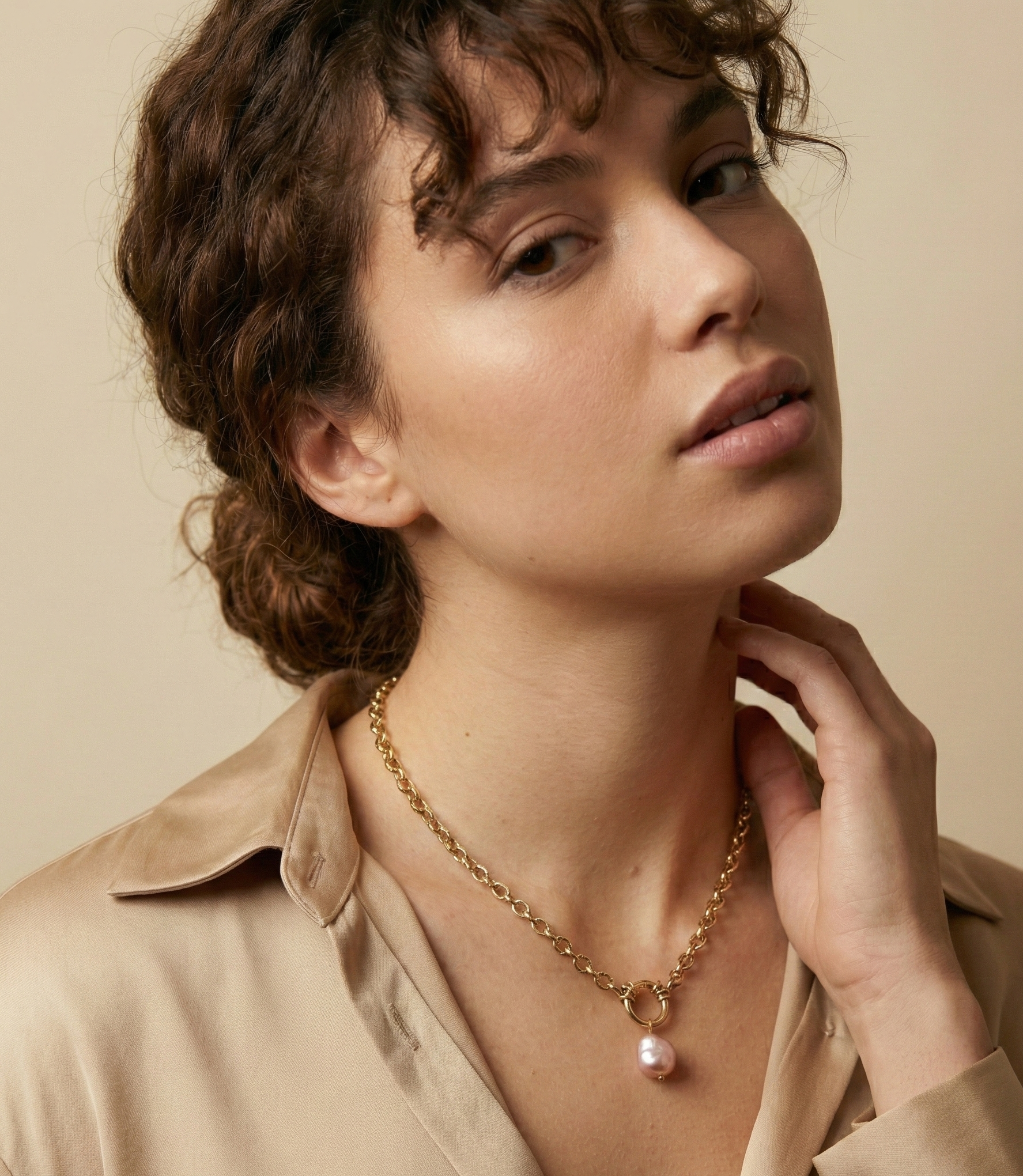 A model with curly brown hair in a bun wearing a beige silk shirt and a Parisian chic gold chain necklace with a pink baroque pearl drop