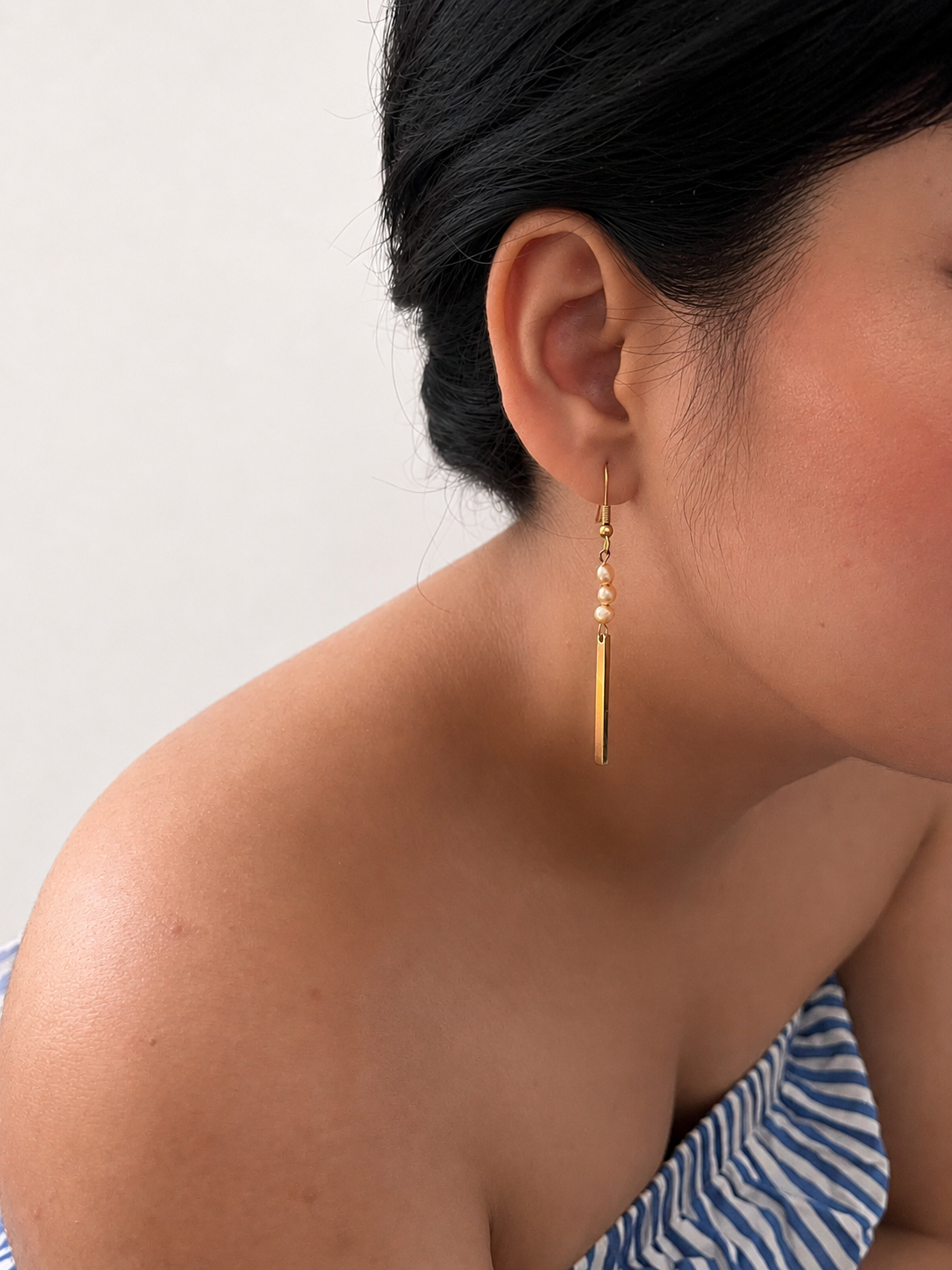 Woman wearing a gold linear drop earring with three petite gold baroque freshwater pearls.