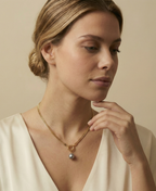 Close-up editorial shot of a woman wearing a minimalist gold chain with a grey baroque pearl drop, highlighting the necklace length and luster against warm skin tones.