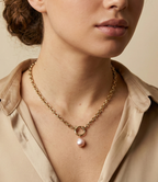 A model with curly brown hair in a bun wearing a beige silk shirt and a Parisian chic gold chain necklace with a pink baroque pearl drop