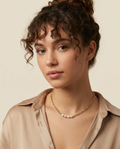 Model with curly brown hair wearing a delicate gold chain necklace with four oval champagne pearls, styled with a beige silk shirt for a Parisian chic look