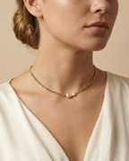 Close-up of a model wearing a dainty gold curb chain necklace featuring a pink baroque pearl and a small champagne pearl, styled with an ivory silk top