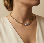 Close-up editorial portrait of a woman wearing a gold Figaro chain necklace featuring a unique grey Keshi baroque pearl with metallic luster. The necklace sits on the collarbone, styled with a deep V-neck cream silk top
