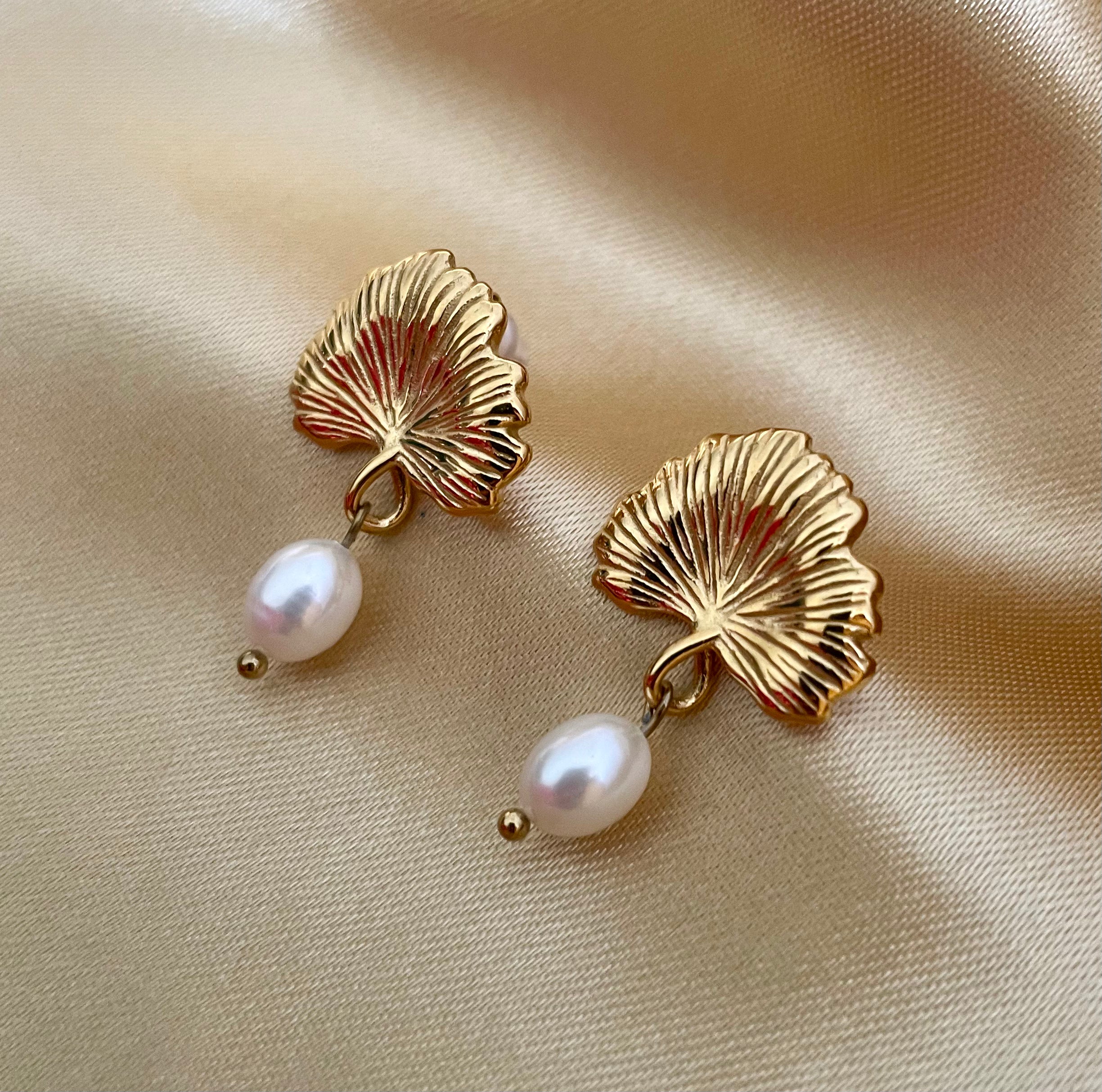 The earrings feature a detailed, textured gold fan shape resembling a ginkgo biloba leaf, from which hangs a lustrous white freshwater pearl.