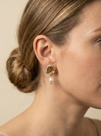 The earrings feature a detailed, textured gold fan shape resembling a ginkgo biloba leaf, from which hangs a lustrous white freshwater pearl. Soft, warm Rembrandt lighting highlights the metallic gold sheen and pearl iridescence against a matte ivory background, showcasing the jewelry's high-end craftsmanship and elegant scale on the ear.