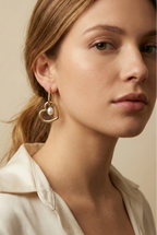 Close-up of a model wearing gold heart-shaped hoop earrings with a lustrous freshwater white pearl charm. High-quality jewelry photography showcasing the polished gold finish and pearl texture against a plain ivory background