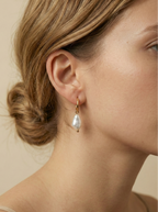 Close-up photography of a dainty gold hoop earring featuring a lustrous white baroque pearl drop worn on a model's ear. High-resolution commercial jewelry shot showcasing organic pearl texture, nacre iridescence, and a simple gold clasp against a matte ivory background.