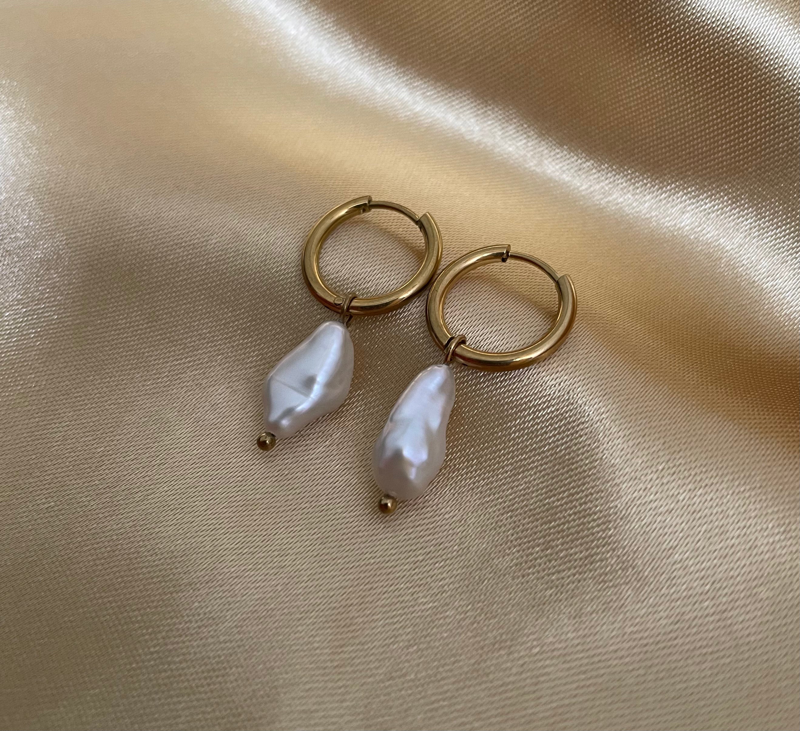 Close-up photography of a dainty gold hoop earring featuring a lustrous white keshi baroque pearl drop. High-resolution commercial jewelry shot showcasing organic pearl texture, nacre iridescence, and a simple gold clasp against a matte ivory background.