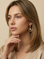 Close-up editorial photography of a gold hoop drop earring featuring a suspended white high-luster pearl, worn by a model with blonde-brown hair against a matte ivory background. High-resolution luxury jewelry shot highlighting realistic gold texture and pearl iridescence.