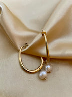 Close-up side profile of a woman's ear wearing a gold hoop earring with a single white pearl drop, highlighting the metallic luster and pearl detail against a soft neutral background.