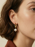 Close-up of a model wearing gold drop earrings featuring a unique knot design and a dangling white round pearl. The earrings are styled with a rust-colored silk shirt
