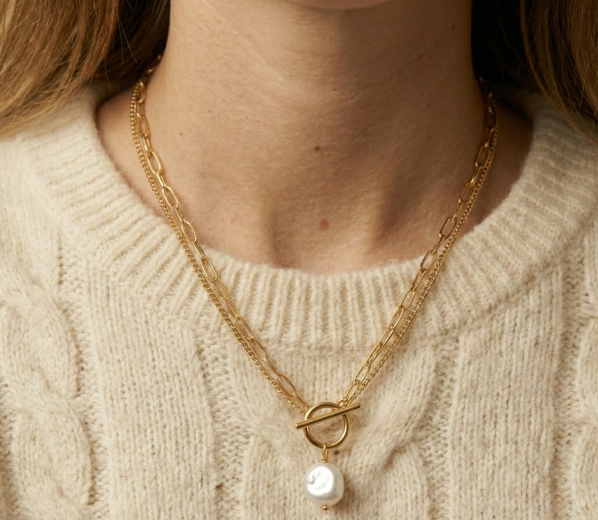 Close-up detail of a double-strand gold chain necklace featuring a lustrous white baroque pearl pendant and a T-bar toggle clasp, worn on the collarbone of a model