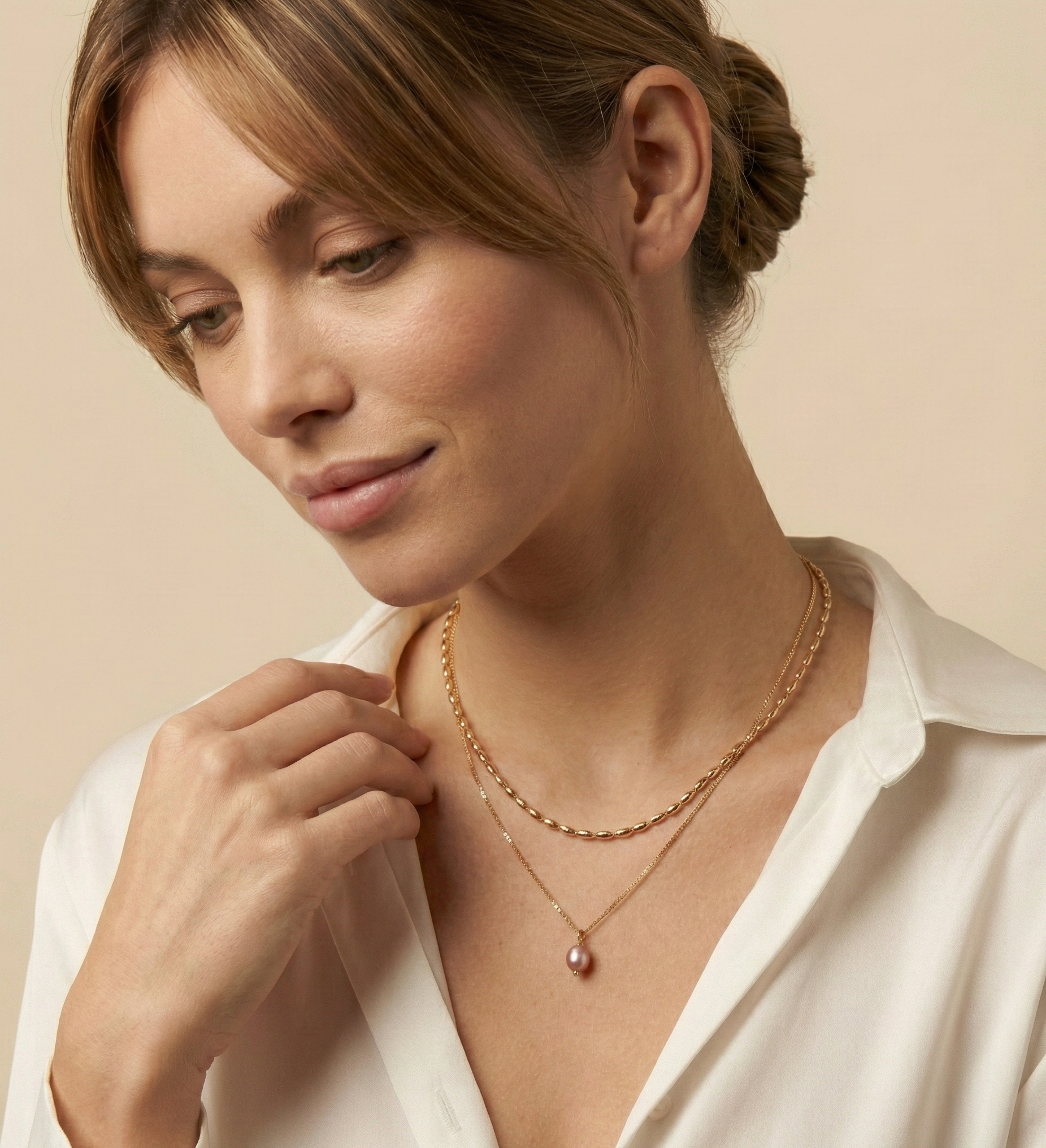 Close-up of a model wearing a layered gold necklace set featuring a choker with small gold beads and a delicate chain with a single dark pink oval pearl pendant, styled with a deep V-neck white silk shirt