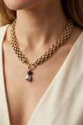 Elegant French model tucking hair behind ear wearing a chunky gold link chain necklace with a T-bar toggle clasp, featuring a drop pendant with a round black pearl and a grey baroque pearl against an ivory top