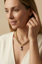 Elegant French model tucking hair behind ear wearing a chunky gold link chain necklace with a T-bar toggle clasp, featuring a drop pendant with a round black pearl and a grey baroque pearl against an ivory top