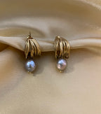 Close-up of 18k gold multi-hoop earrings featuring a lustrous grey pearl drop. The jewelry highlights a textured gold finish and organic pearl shape, set against a plain ivory background