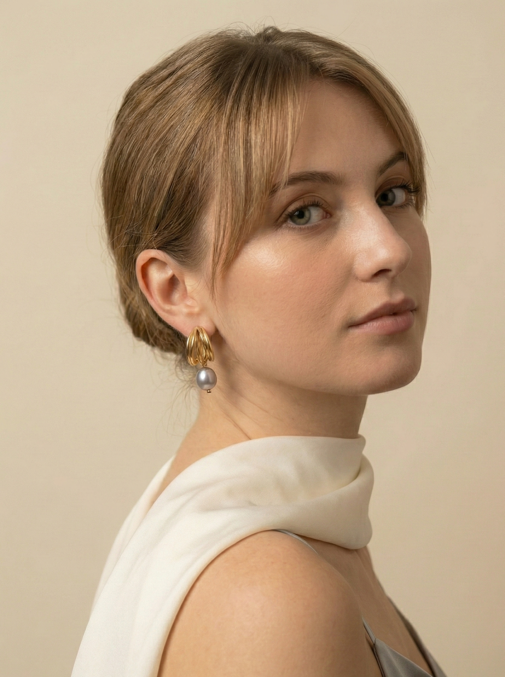 Close-up of 18k gold multi-hoop earrings featuring a lustrous grey pearl drop, worn by a model with a low bun hairstyle. The jewelry highlights a textured gold finish and organic pearl shape, set against a plain ivory background