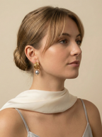 Close-up of 18k gold multi-hoop earrings featuring a lustrous grey pearl drop, worn by a model with a low bun hairstyle. The jewelry highlights a textured gold finish and organic pearl shape, set against a plain ivory background.