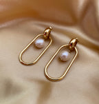Close-up view of gold elongated oval hoop earrings featuring a suspended white baroque pearl, shown worn on the ear to verify size and scale