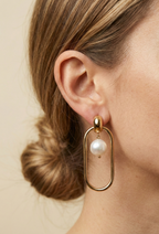 Close-up view of gold elongated oval hoop earrings featuring a suspended white baroque pearl