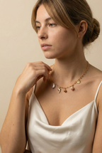 Model wearing a gold paperclip chain necklace featuring a white keshi pearl, gold dolphin charm, pink baroque pearl, and gold sunflower charm, styled with a white silk camisole