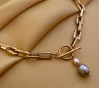 Detail shot of a luxury gold link necklace worn on an ivory silk top. The focal point is a T-bar toggle closure holding a high-shine metallic grey baroque pearl and a champagne oval pearl, showing deep luster and smooth nacre