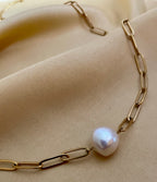 Close-up of a model wearing a gold paperclip chain necklace featuring a single white baroque pearl pendant, styled with a cream silk deep V-neck top