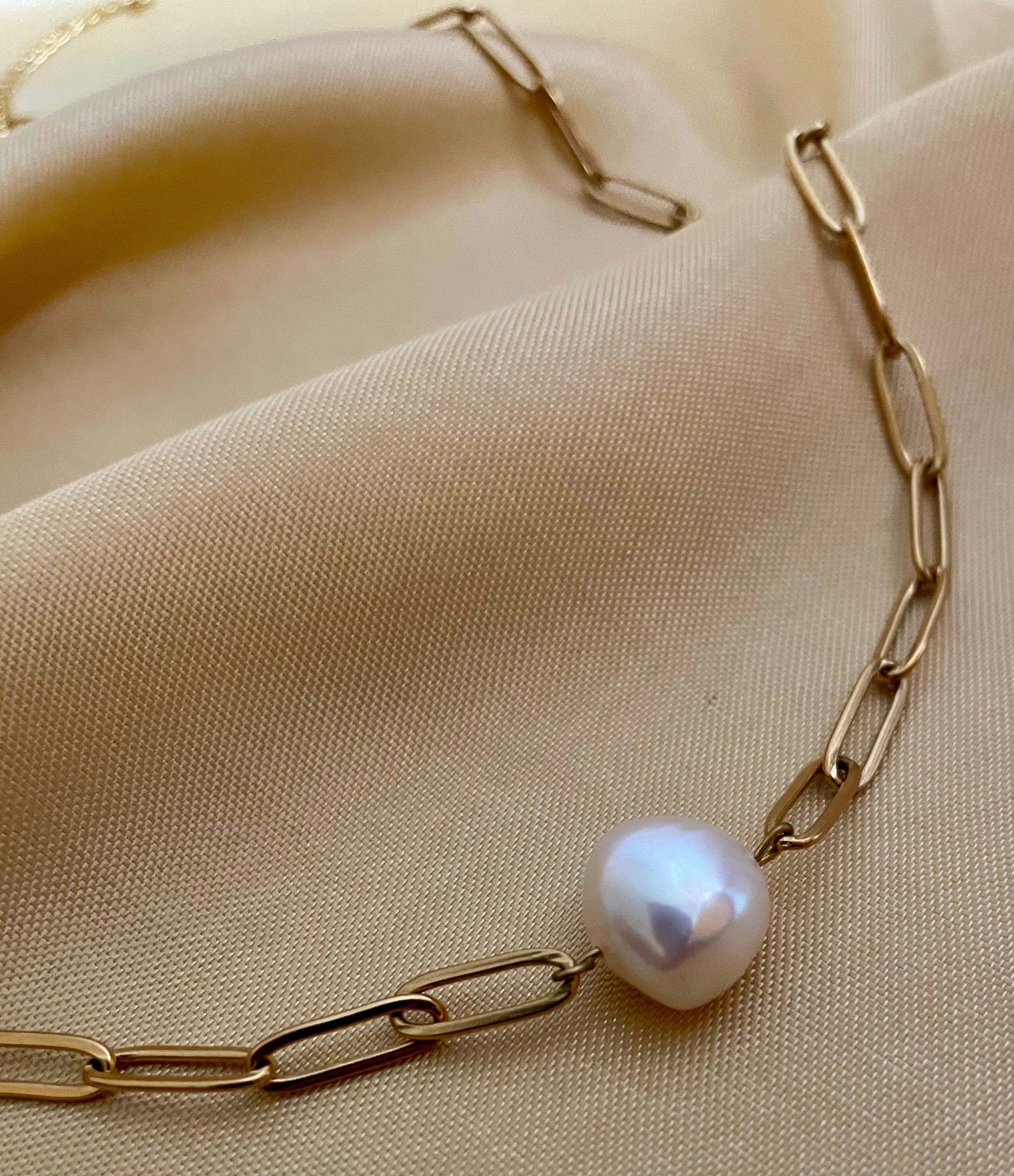 Close-up of a model wearing a gold paperclip chain necklace featuring a single white baroque pearl pendant, styled with a cream silk deep V-neck top