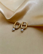 Close-up of gold rectangular huggie hoop earrings featuring a dainty grey pearl drop charm, worn on the ear.
