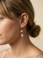 Close-up detail of gold scallop shell earrings featuring a lustrous white pearl drop. The earrings are worn by a model to show scale, highlighting the textured gold finish and elegant dangle design perfect for beach weddings or summer luxury