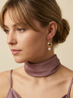 French style inspiration: A model with blonde-brown hair in a messy low bun wearing textured gold sea shell huggie earrings with a dainty pearl charm. Soft studio lighting highlights the iridescence of the pearl and the model's mauve camisole