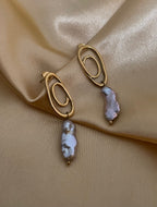 Close-up of a  luxury grey baroque keshi pearl drop earrings with a sculptural gold double-oval link stud. The image highlights the iridescent nacre and organic shape of the pearl, styled against a soft matte ivory background, representing high-end commercial jewelry photography