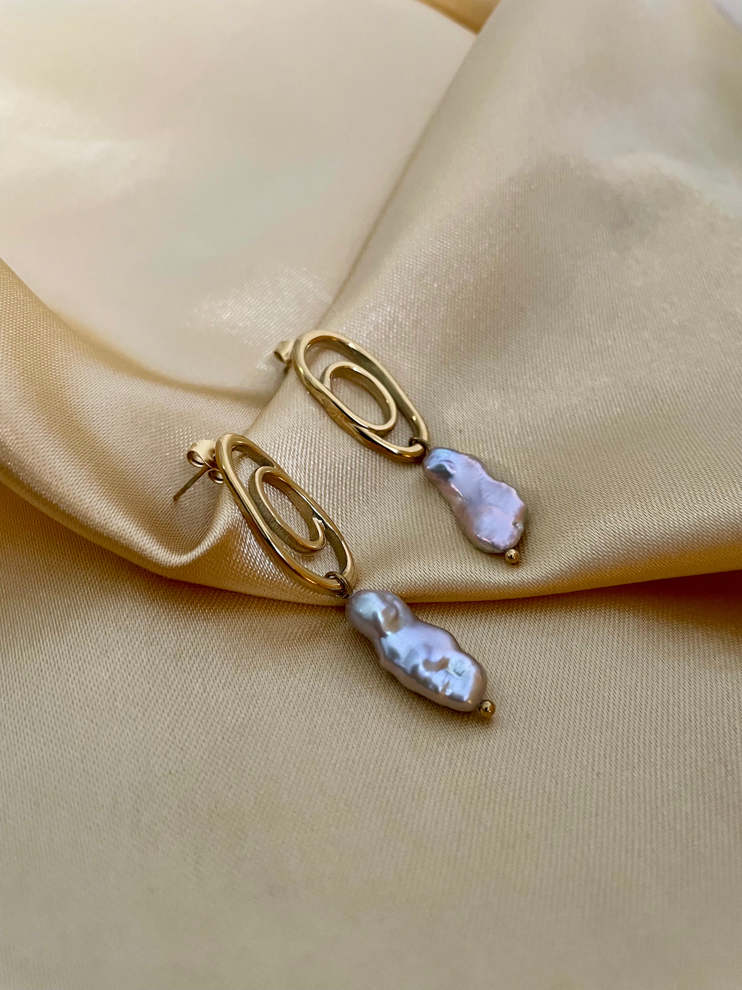 Close-up of a  luxury grey baroque keshi pearl drop earrings with a sculptural gold double-oval link stud. The image highlights the iridescent nacre and organic shape of the pearl, styled against a soft matte ivory background, representing high-end commercial jewelry photography