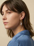Close-up profile of a french model wearing luxury grey baroque pearl drop earrings with a sculptural gold double-oval link stud. The image highlights the iridescent nacre and organic shape of the pearl, styled with a blue knit top against a soft matte ivory background, representing high-end commercial jewelry photography