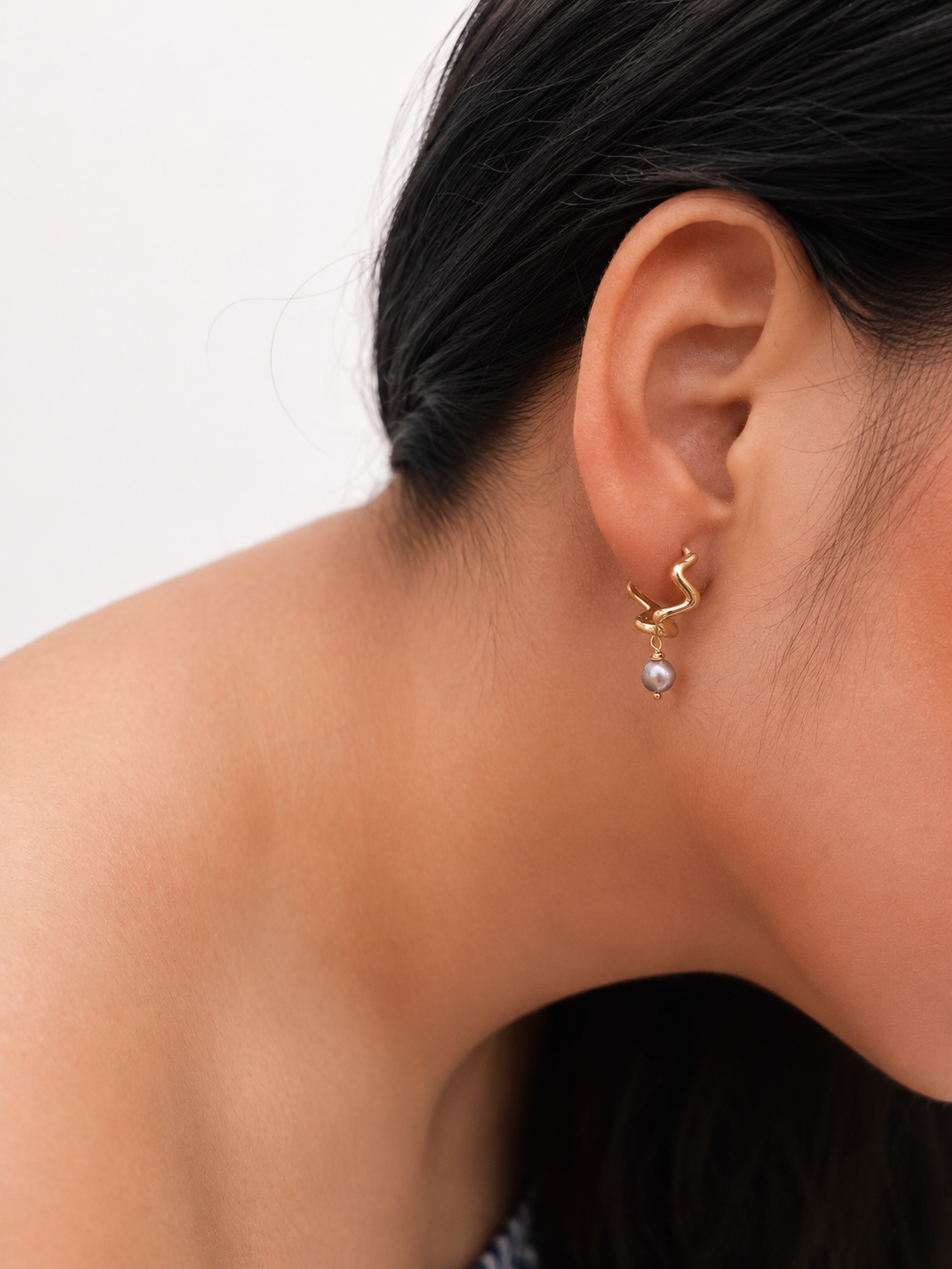 Close-up of a gold Waves petit creole earring with a grey baroque freshwater pearl worn on the ear.